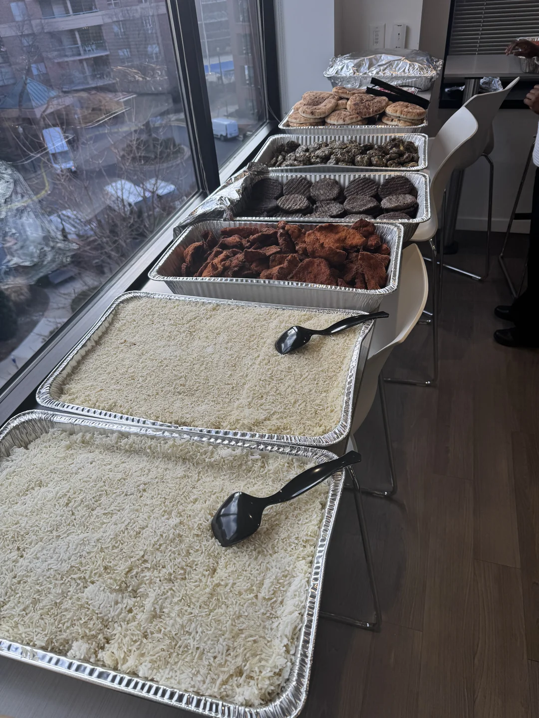 Catering spread of foil trays with rice, crispy falafel patties, fried eggplant and pita breads by a window
