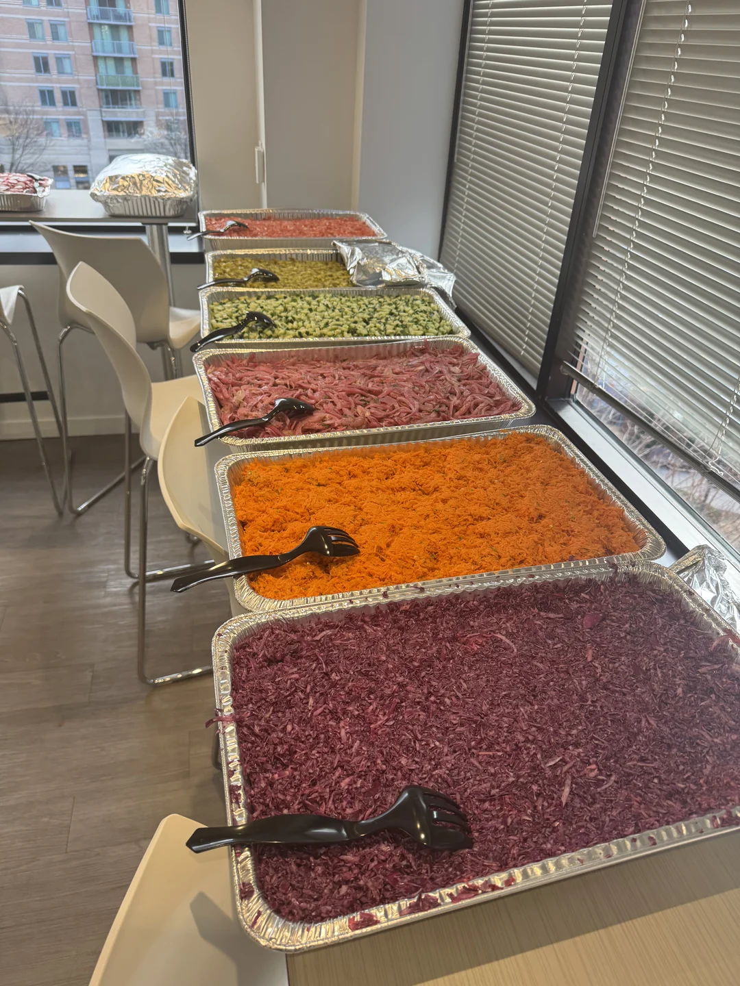 Catering spread of foil trays filled with shredded carrot, red cabbage, pickled salads and olives by a window