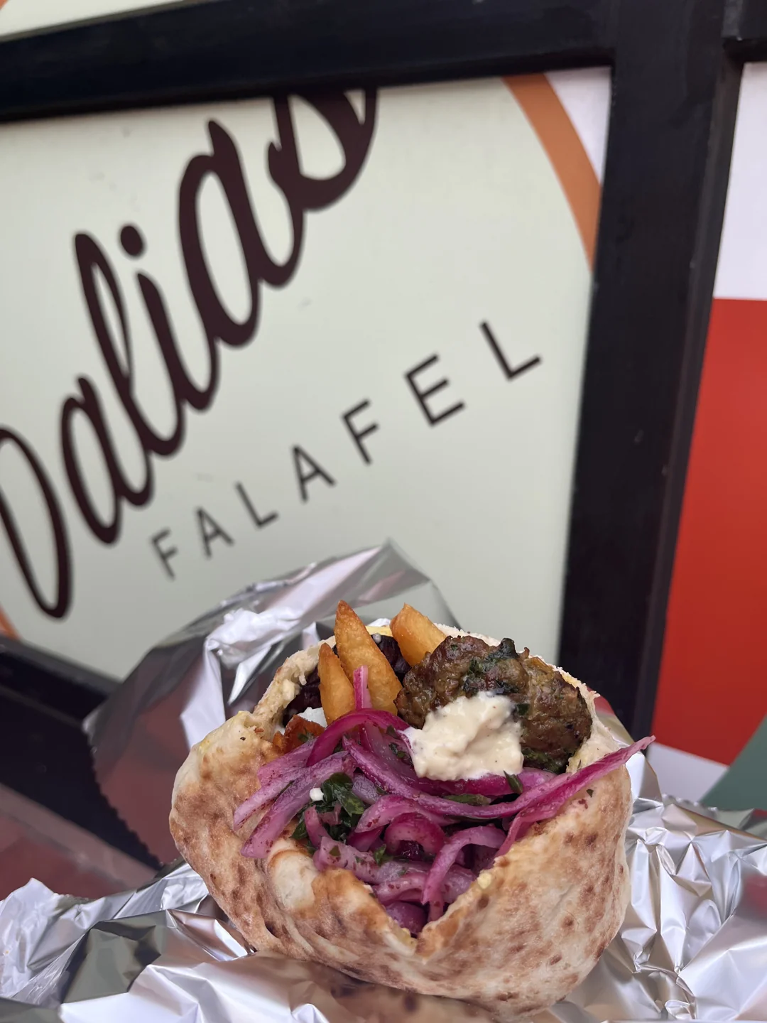 Stuffed pita with kabob, fries and pickled red onions in front of the Dalia's Falafel sign